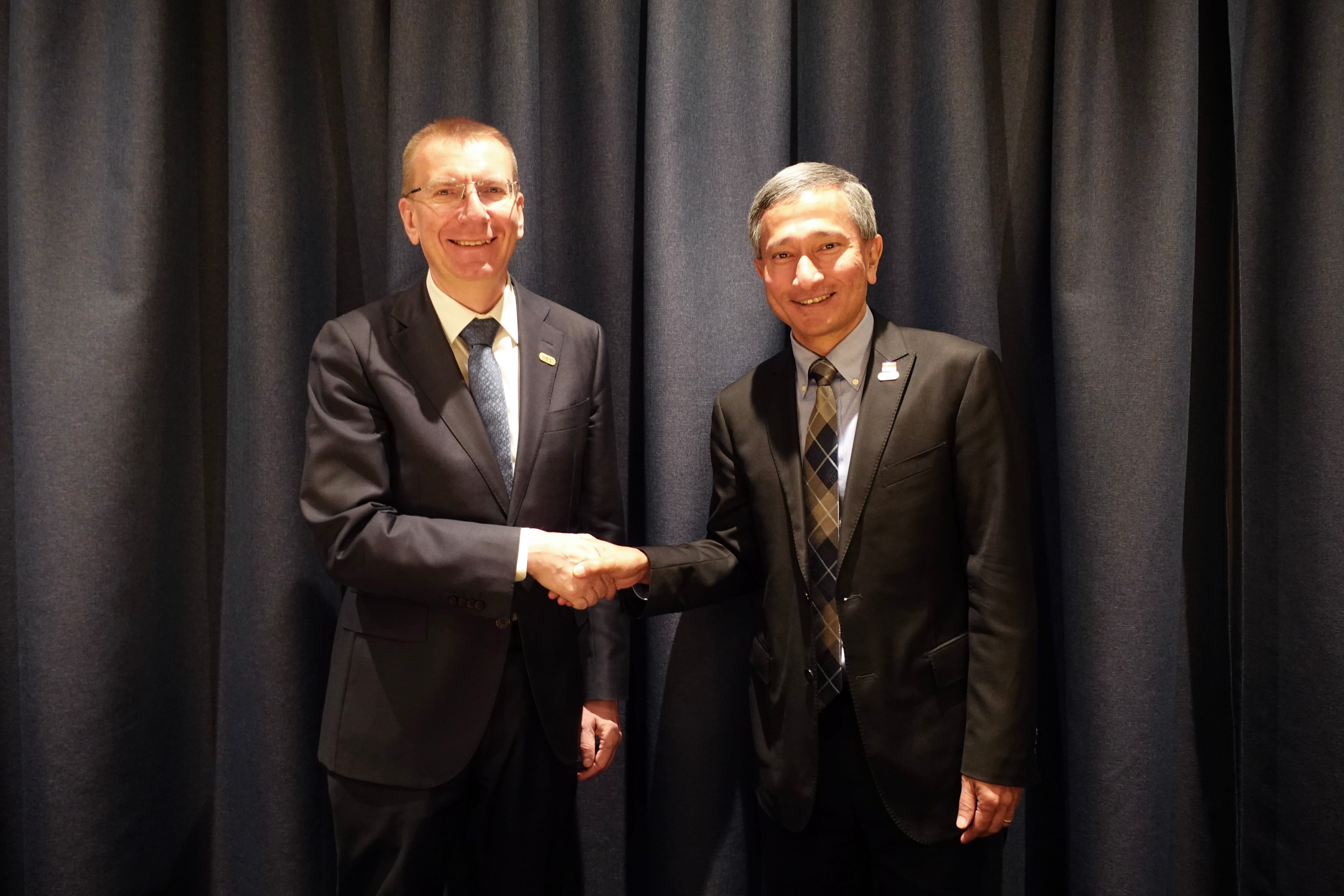Two men in suits shaking hands in front of a blue curtain backdrop.